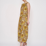 A woman modeling a sleeveless, floor-length dress with a bold yellow, gold, and brown floral pattern against a plain white studio background.