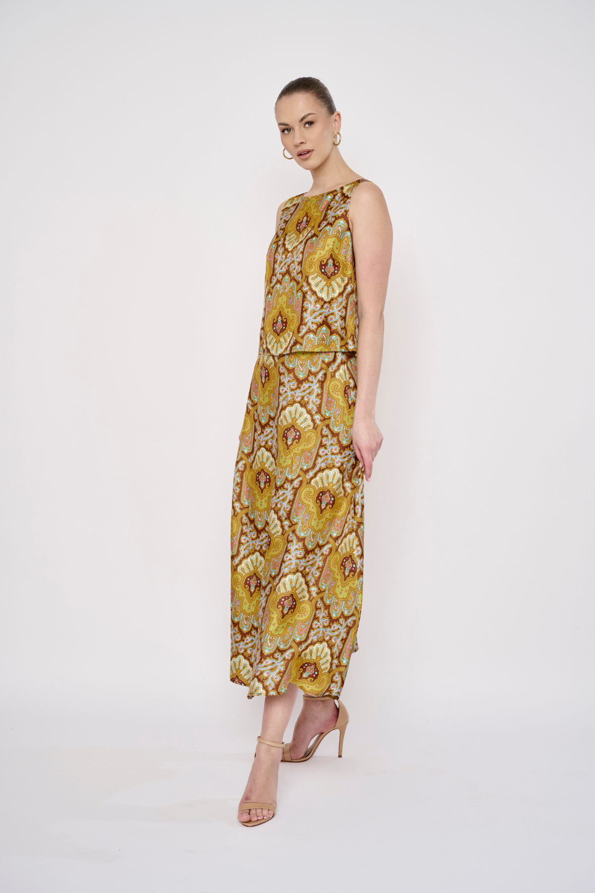 A woman modeling a sleeveless, floor-length dress with a bold yellow, gold, and brown floral pattern against a plain white studio background.