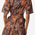 A woman wearing a short-sleeve collared dress with a tiered ruffled skirt, featuring a navy blue and rust-orange floral pattern. The photo is taken from the back against a plain light gray background.