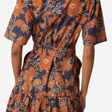 A woman wearing a short-sleeve collared dress with a tiered ruffled skirt, featuring a navy blue and rust-orange floral pattern. The photo is taken from the back against a plain light gray background.