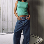 A woman stands in front of a gray wood-paneled wall wearing a mint green sleeveless crop top and wide-leg blue jeans, with a white surfboard behind her.