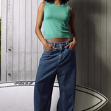 A woman stands in front of a gray wood-paneled wall wearing a mint green sleeveless crop top and wide-leg blue jeans, with a white surfboard behind her.