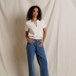 A woman stands against a draped cream fabric backdrop wearing blue wide-leg jeans, a white short-sleeve henley-style top, and dark brown shoes.