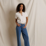 A woman stands against a draped cream fabric backdrop wearing blue wide-leg jeans, a white short-sleeve henley-style top, and dark brown shoes.