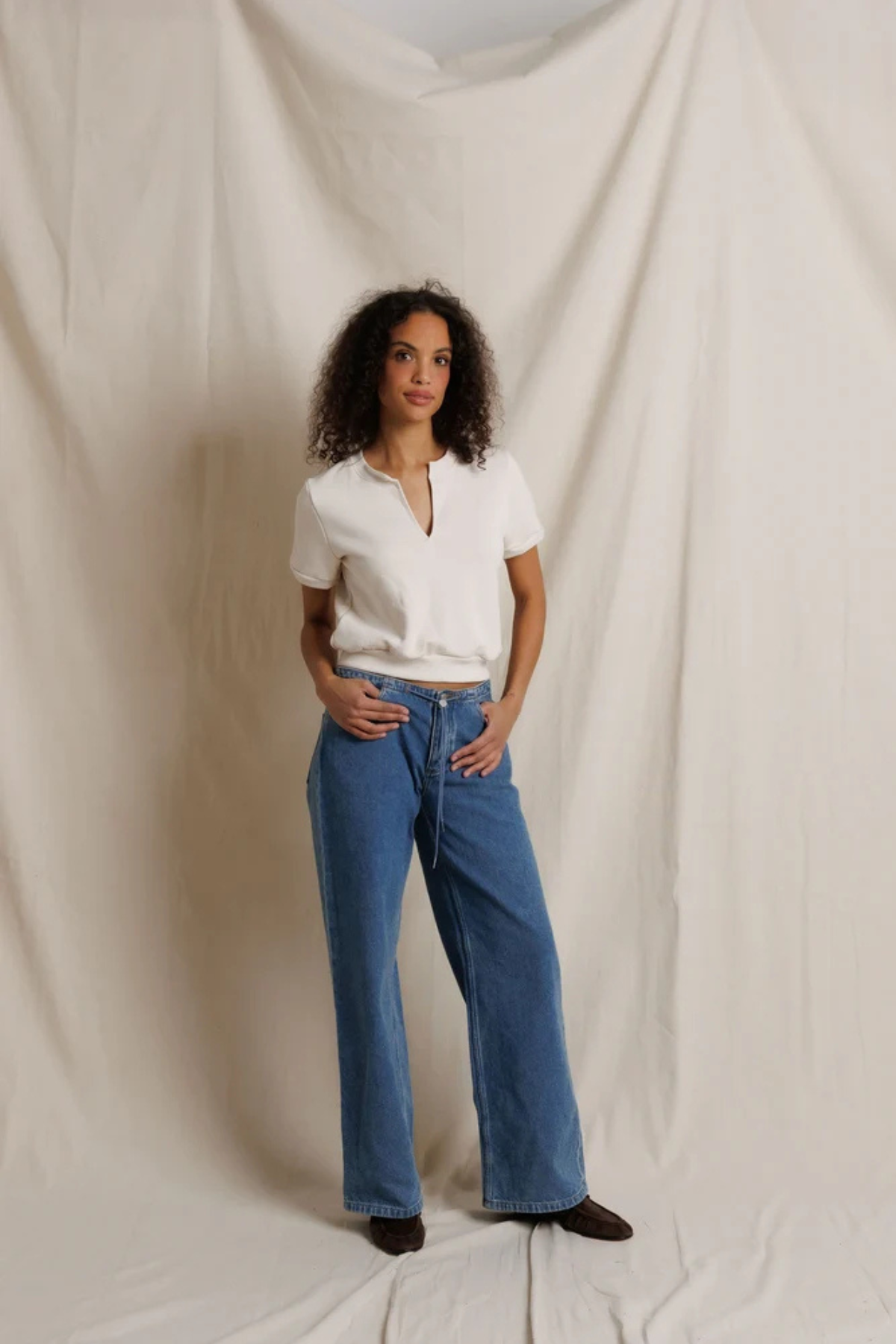 A woman stands against a draped cream fabric backdrop wearing blue wide-leg jeans, a white short-sleeve henley-style top, and dark brown shoes.