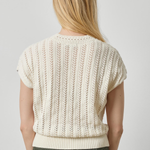 A woman viewed from the back wearing a cream-colored short-sleeve knit sweater with vertical textured stripes and olive green pants, set against a plain light gray studio background.