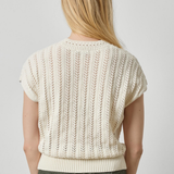 A woman viewed from the back wearing a cream-colored short-sleeve knit sweater with vertical textured stripes and olive green pants, set against a plain light gray studio background.