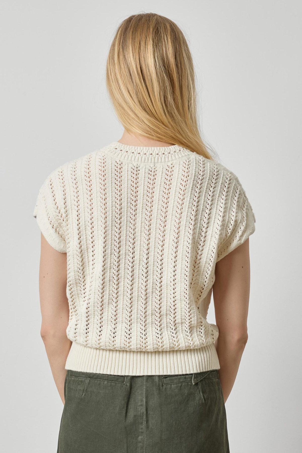 A woman viewed from the back wearing a cream-colored short-sleeve knit sweater with vertical textured stripes and olive green pants, set against a plain light gray studio background.