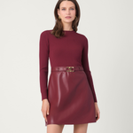 A woman modeling a ribbed long-sleeve knit top and a shiny faux-leather mini skirt in a matching deep burgundy color, accessorized with a matching belt and a gold buckle, against a plain light gray studio background.