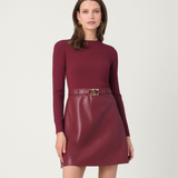 A woman modeling a ribbed long-sleeve knit top and a shiny faux-leather mini skirt in a matching deep burgundy color, accessorized with a matching belt and a gold buckle, against a plain light gray studio background.