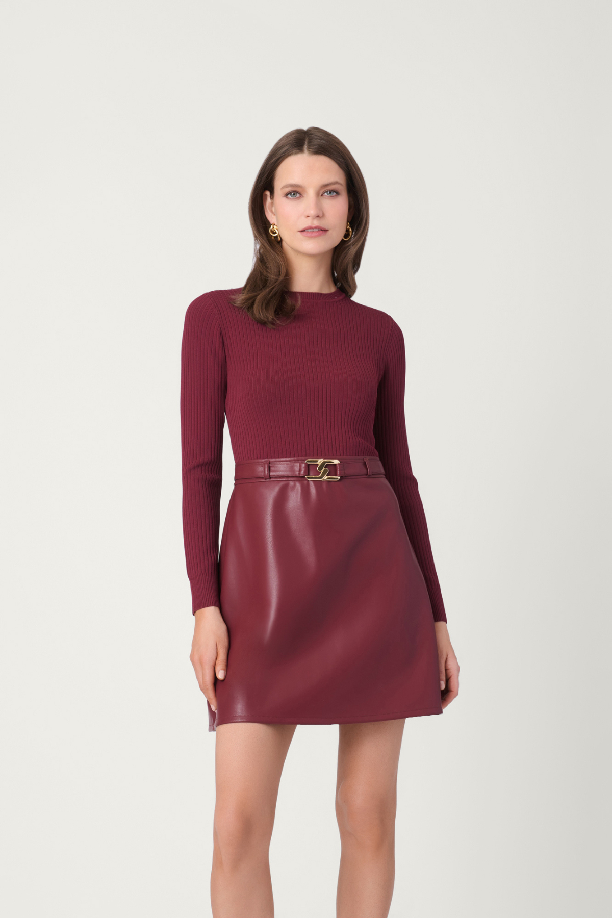 A woman modeling a ribbed long-sleeve knit top and a shiny faux-leather mini skirt in a matching deep burgundy color, accessorized with a matching belt and a gold buckle, against a plain light gray studio background.