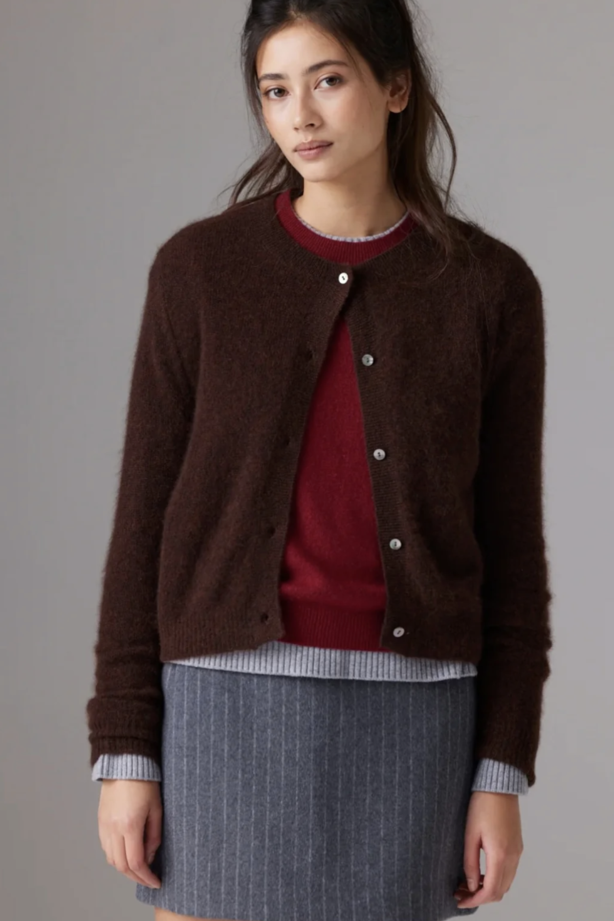 A woman wearing a dark brown fuzzy button-up cardigan over a burgundy sweater and a gray pinstriped skirt, photographed against a neutral gray studio background.