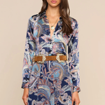 A woman modeling a long-sleeve button-front dress with a blue, beige, and white paisley pattern, styled with a tan belt with silver buckle, against a solid beige background.