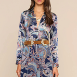 A woman modeling a long-sleeve button-front dress with a blue, beige, and white paisley pattern, styled with a tan belt with silver buckle, against a solid beige background.