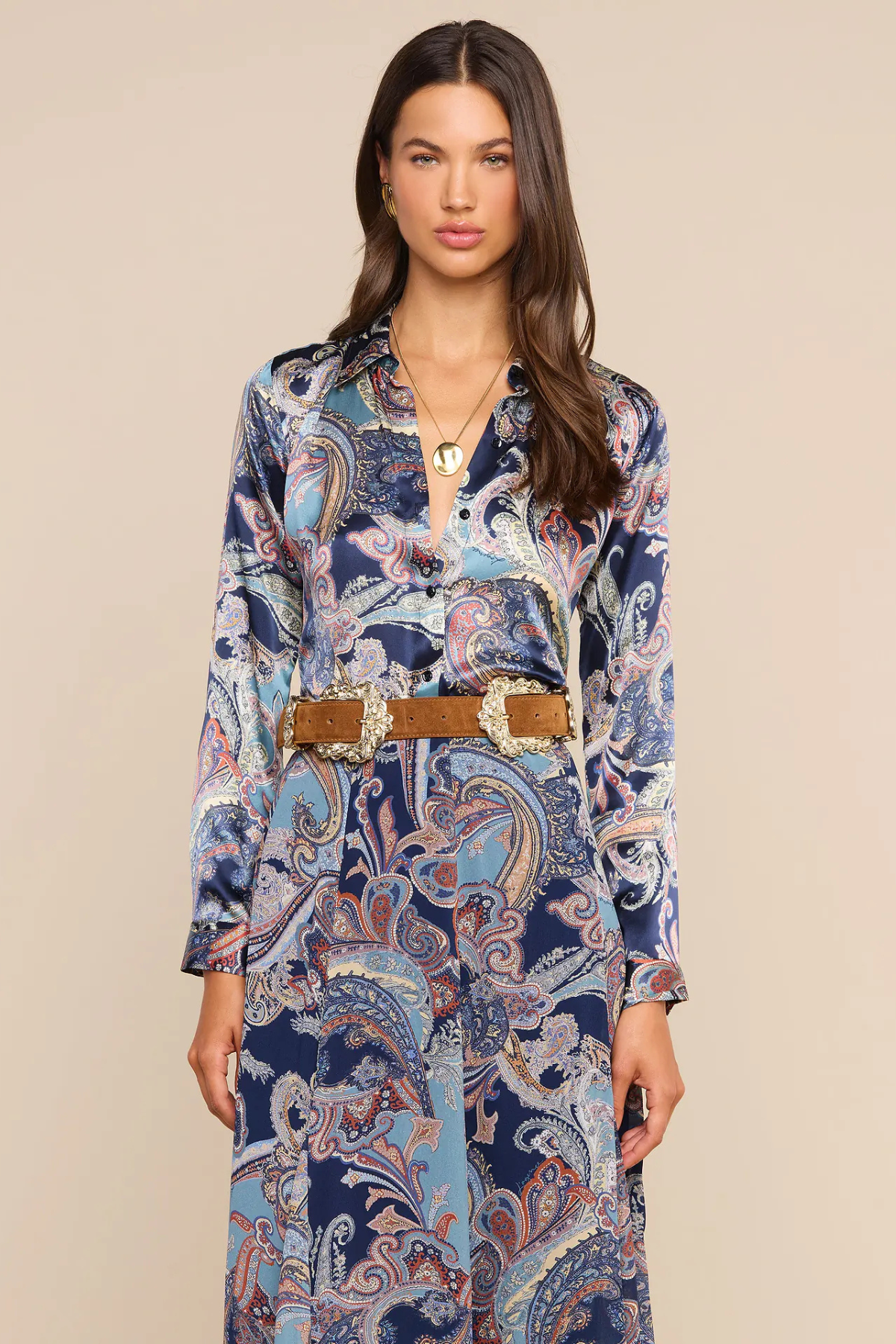 A woman modeling a long-sleeve button-front dress with a blue, beige, and white paisley pattern, styled with a tan belt with silver buckle, against a solid beige background.