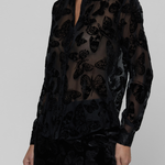 A model wearing a sheer black long-sleeve button-up shirt with a 3D butterfly pattern, paired with matching black trousers, against a solid gray studio background.
