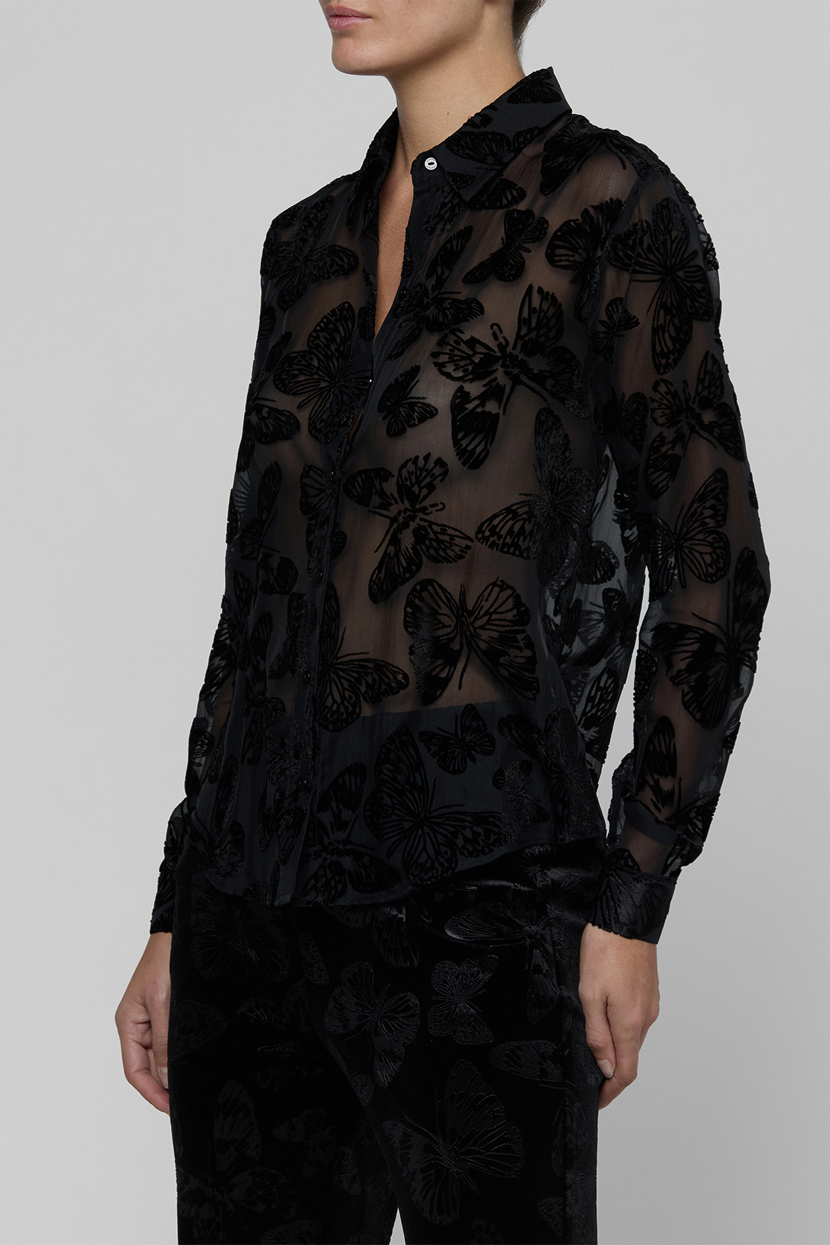 A model wearing a sheer black long-sleeve button-up shirt with a 3D butterfly pattern, paired with matching black trousers, against a solid gray studio background.