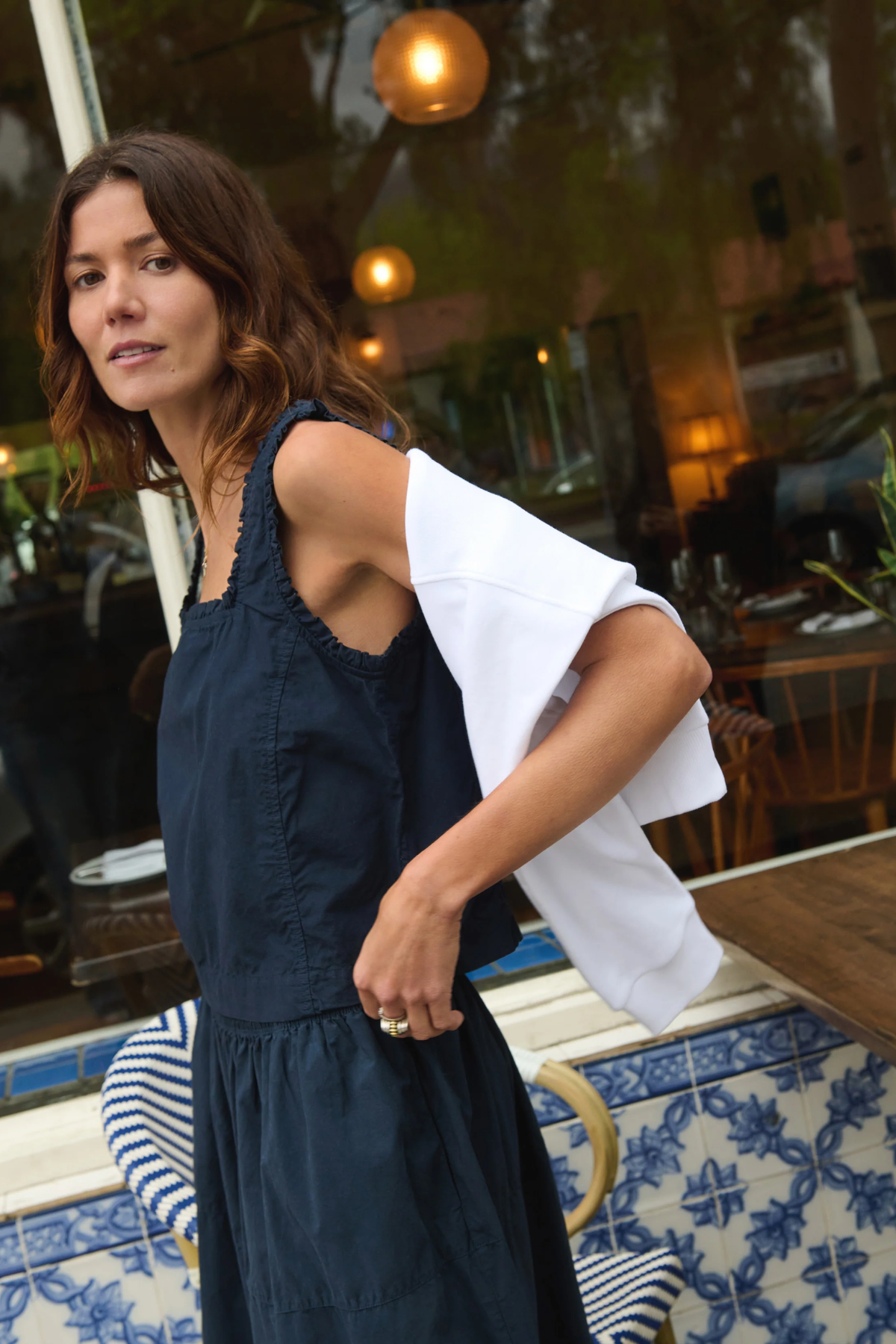 Woman in a dark blue dress with a white shirt over her shoulder, standing on a tiled patio.