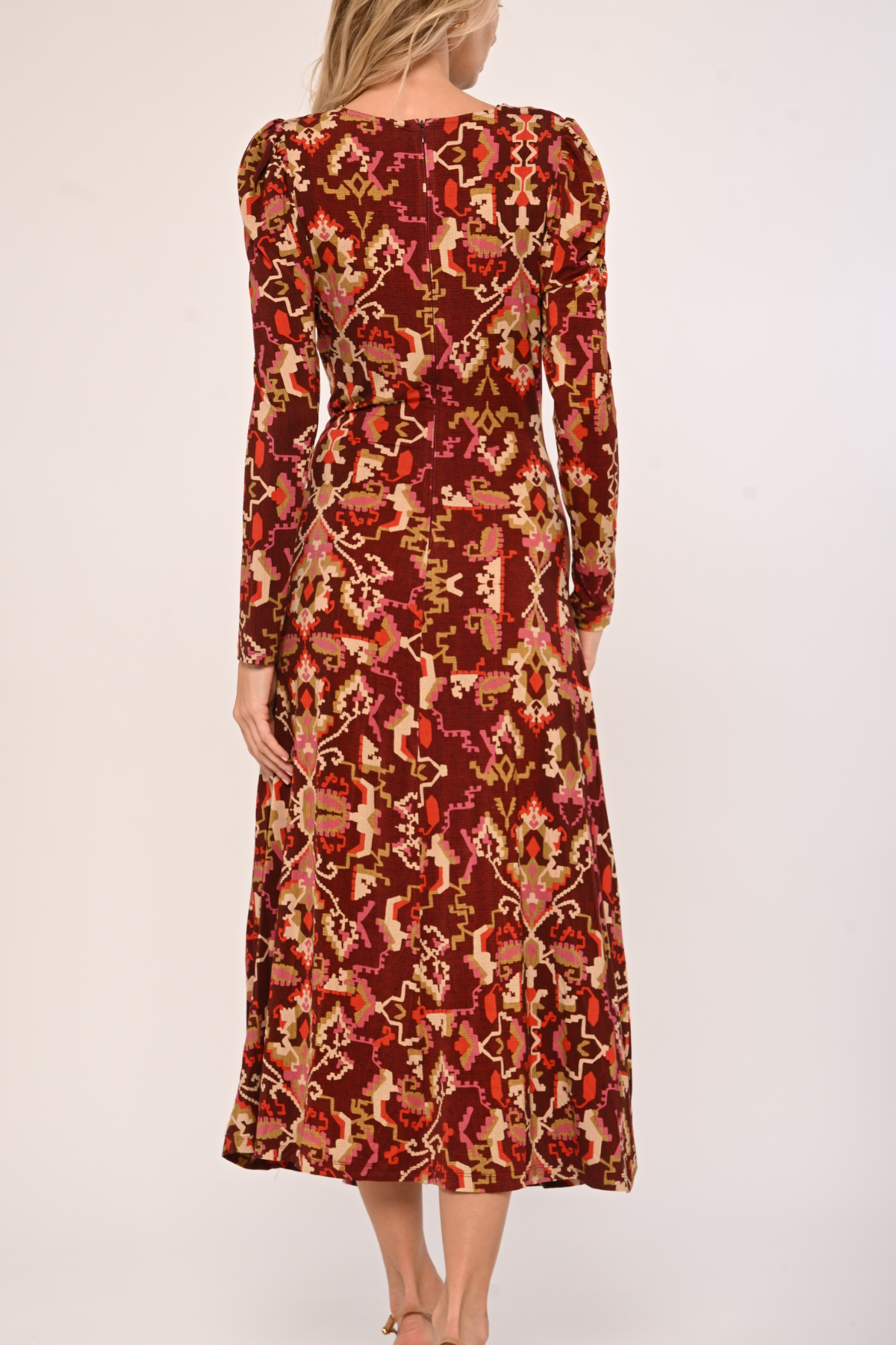 A woman modeling a long-sleeve midi dress with a red, orange, and beige floral ikat pattern, shown from the back against a plain light background.