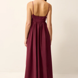 A woman wearing a long, sleeveless maxi dress in a deep burgundy or plum color, shown from the back against a plain off-white studio background.