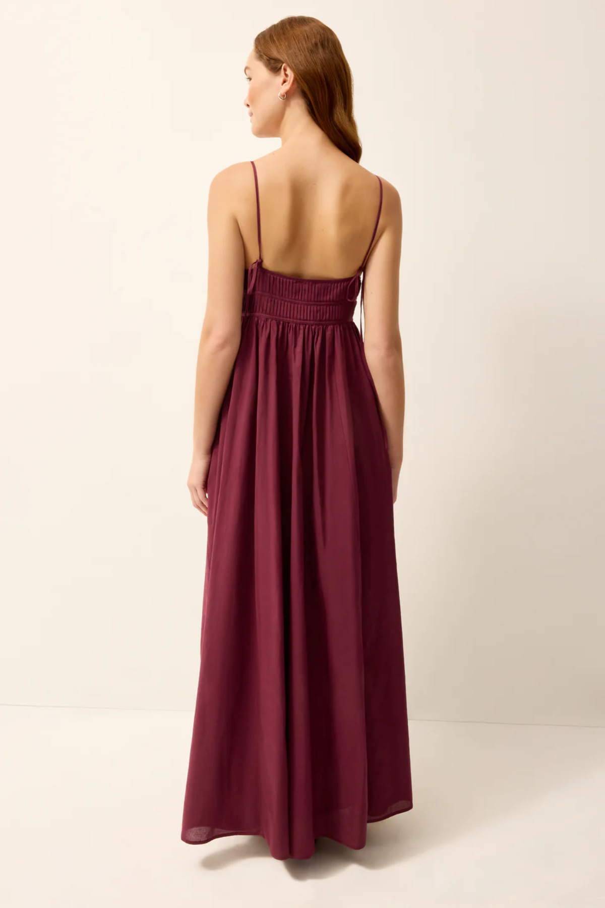 A woman wearing a long, sleeveless maxi dress in a deep burgundy or plum color, shown from the back against a plain off-white studio background.