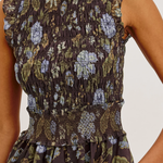 Close-up of a sleeveless smocked dress with a dark brown base and a muted floral print in olive green, lavender, and beige, featuring ruffled cap sleeves and a smocked waist.