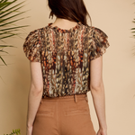 A woman shown from the back wearing a short-sleeve blouse with a brown, beige, and orange abstract pattern, paired with brown pants, set against a warm beige background with decorative plants.
