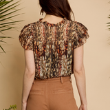 A woman shown from the back wearing a short-sleeve blouse with a brown, beige, and orange abstract pattern, paired with brown pants, set against a warm beige background with decorative plants.