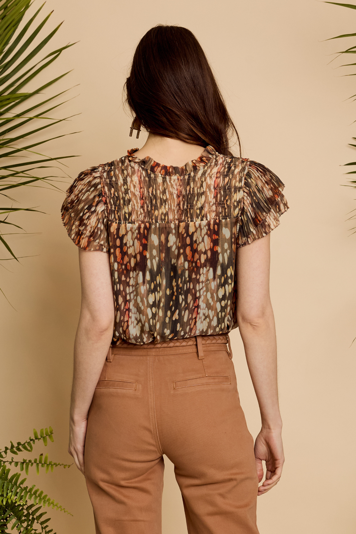 A woman shown from the back wearing a short-sleeve blouse with a brown, beige, and orange abstract pattern, paired with brown pants, set against a warm beige background with decorative plants.