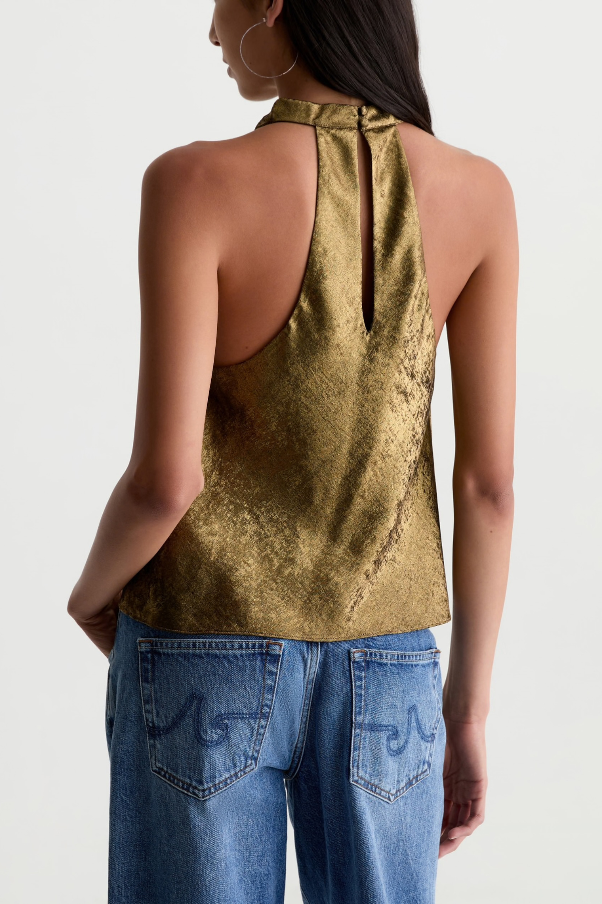 A woman shown from the back wearing a metallic gold halter top and blue jeans against a plain light gray studio background.