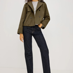 A model wearing an olive green cropped double-breasted jacket, dark wash straight-leg jeans, a light gray t-shirt, and black heeled sandals, standing against a plain light gray studio background.