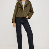 A model wearing an olive green cropped double-breasted jacket, dark wash straight-leg jeans, a light gray t-shirt, and black heeled sandals, standing against a plain light gray studio background.