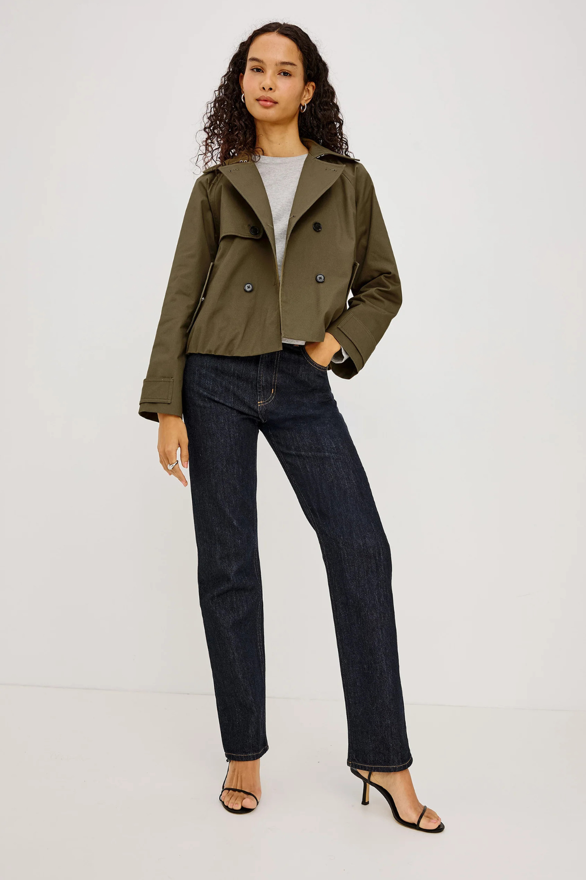 A model wearing an olive green cropped double-breasted jacket, dark wash straight-leg jeans, a light gray t-shirt, and black heeled sandals, standing against a plain light gray studio background.