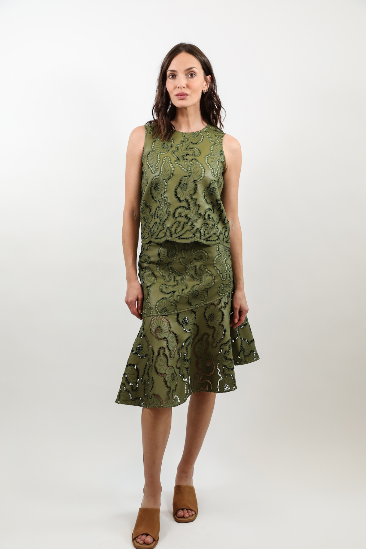 A woman modeling a sleeveless olive green midi dress with a textured, patterned surface and a ruffled hem, paired with brown slide sandals, against a plain light gray studio background.