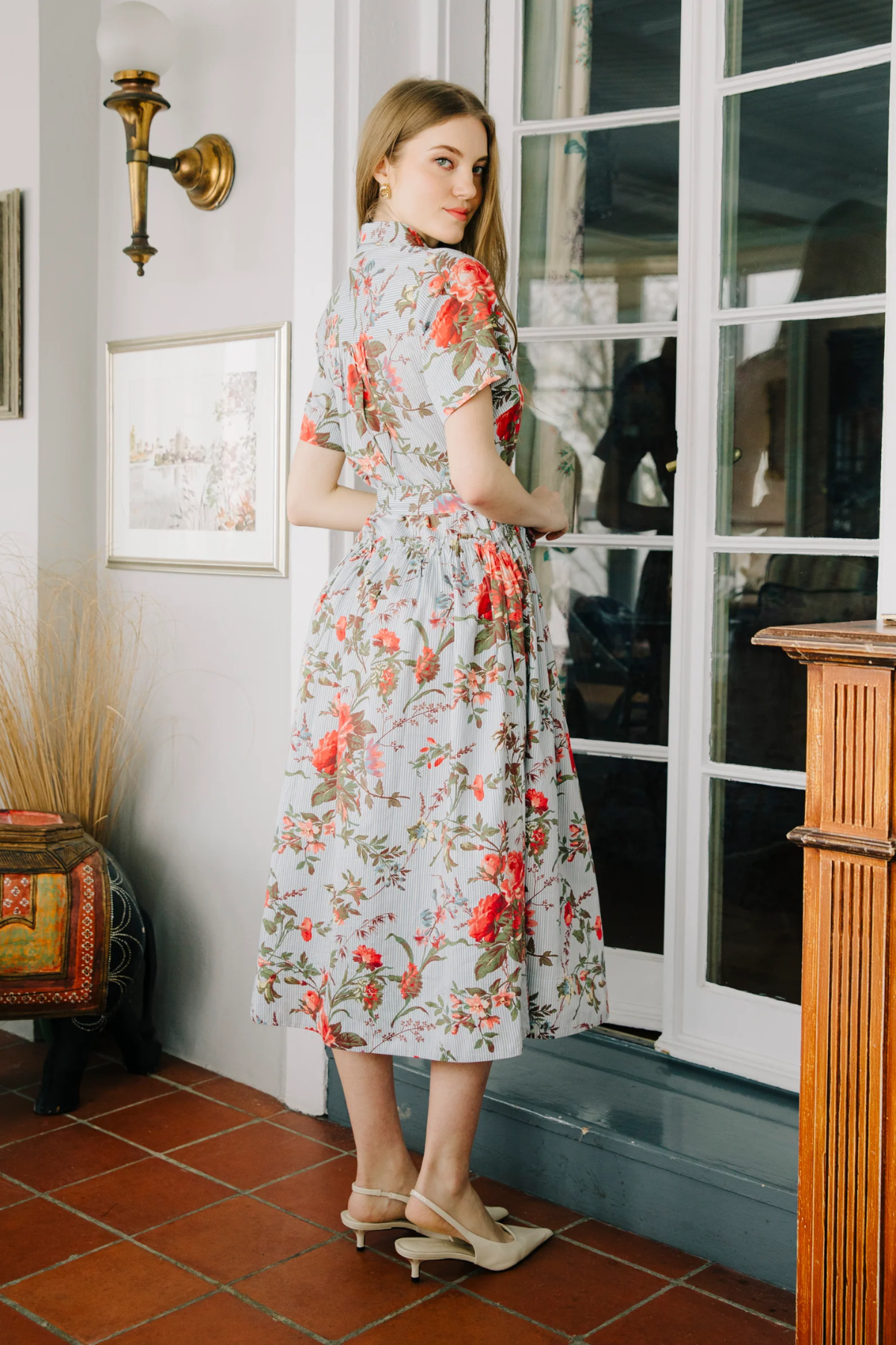 Woman in a floral dress standing in a room with a tiled floor and framed pictures on the wall.