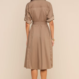 A woman viewed from the back wearing a taupe-brown short-sleeve collared shirt dress with a matching waist tie, paired with beige high-heeled sandals, against a solid beige studio background.