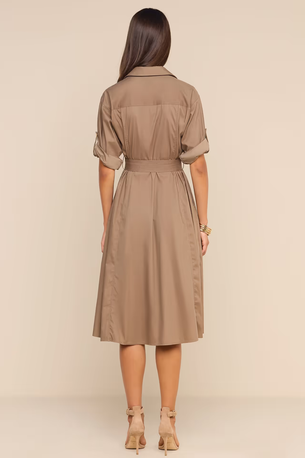 A woman viewed from the back wearing a taupe-brown short-sleeve collared shirt dress with a matching waist tie, paired with beige high-heeled sandals, against a solid beige studio background.