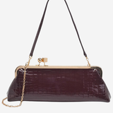 A dark plum purple crocodile-embossed clutch handbag with a gold-tone kiss-lock closure, a thin black shoulder strap, and a gold chain link accent, shown against a plain white background.
