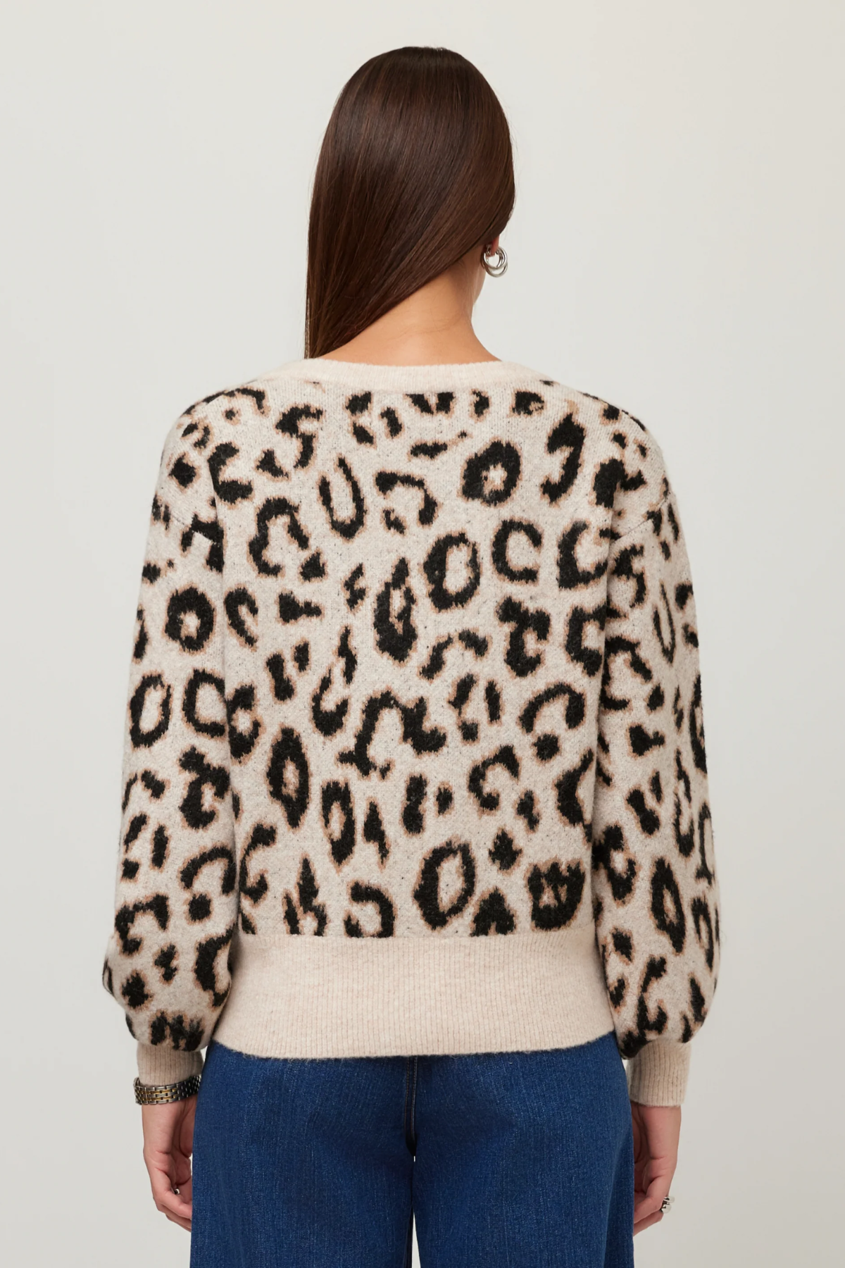 A woman viewed from the back wearing a cream-colored knit sweater with a black leopard print pattern and blue jeans, set against a plain light gray background.