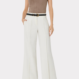 A model wearing a sleeveless taupe-brown V-neck top, white wide-leg trousers with a thin black belt, and black pointed shoes, photographed against a light gray studio background.