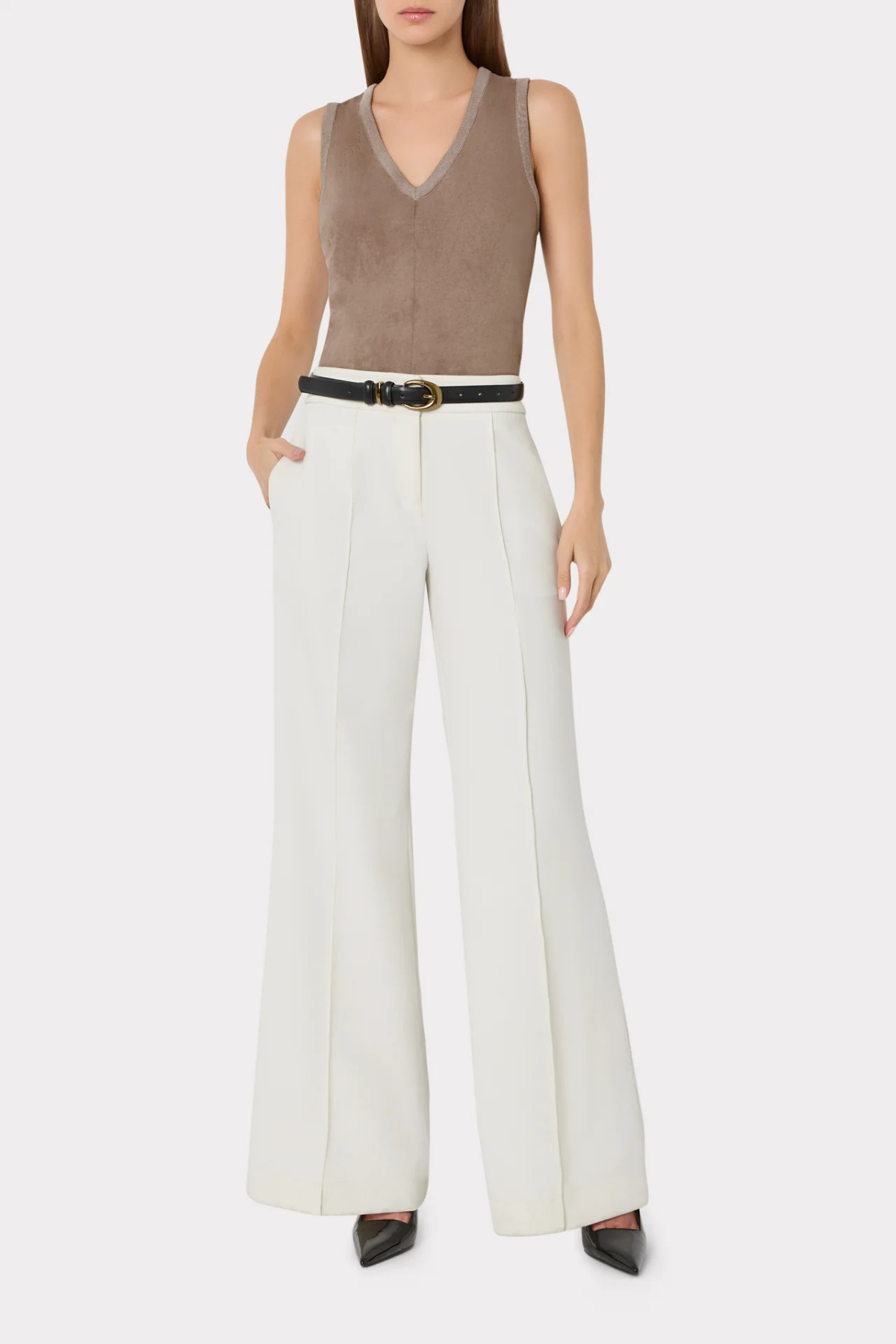 A model wearing a sleeveless taupe-brown V-neck top, white wide-leg trousers with a thin black belt, and black pointed shoes, photographed against a light gray studio background.