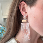 A close-up of a woman wearing a long, dangling statement earring. The earring features a gold-toned geometric setting with a small rectangular stone and a large, cascading fringe tassel made of clear and light pink beads.