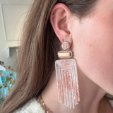 A close-up of a woman wearing a long, dangling statement earring. The earring features a gold-toned geometric setting with a small rectangular stone and a large, cascading fringe tassel made of clear and light pink beads.