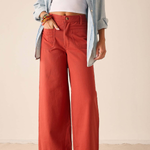 A model wearing rust-colored wide-leg pants, styled with a light blue button-up shirt, a white crop top, and white flip-flop sandals against a neutral beige background.