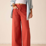 A model wearing rust-colored wide-leg pants, styled with a light blue button-up shirt, a white crop top, and white flip-flop sandals against a neutral beige background.