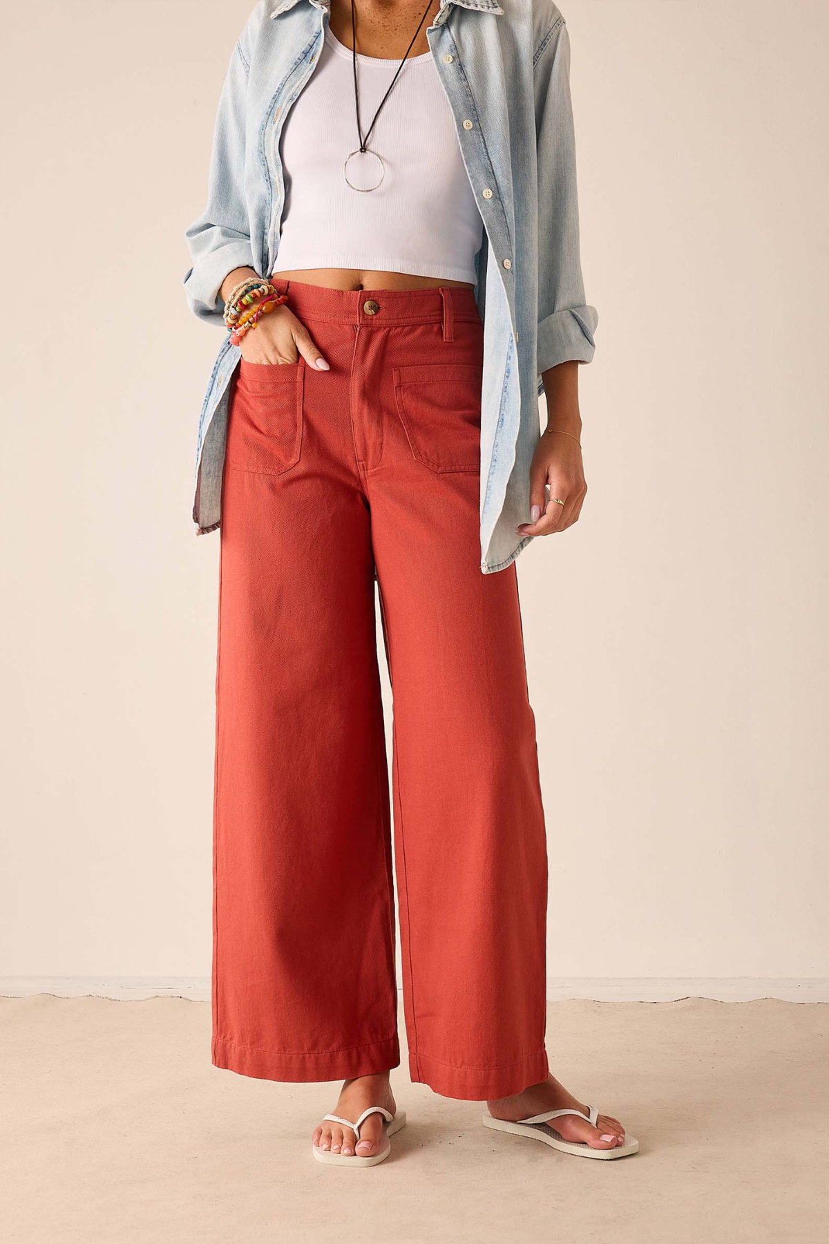 A model wearing rust-colored wide-leg pants, styled with a light blue button-up shirt, a white crop top, and white flip-flop sandals against a neutral beige background.