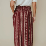 A person shown from the back wearing a white blouse and a high-waisted, pleated maxi skirt with vertical burgundy and beige leafy patterns, paired with brown heeled sandals against a solid beige background.