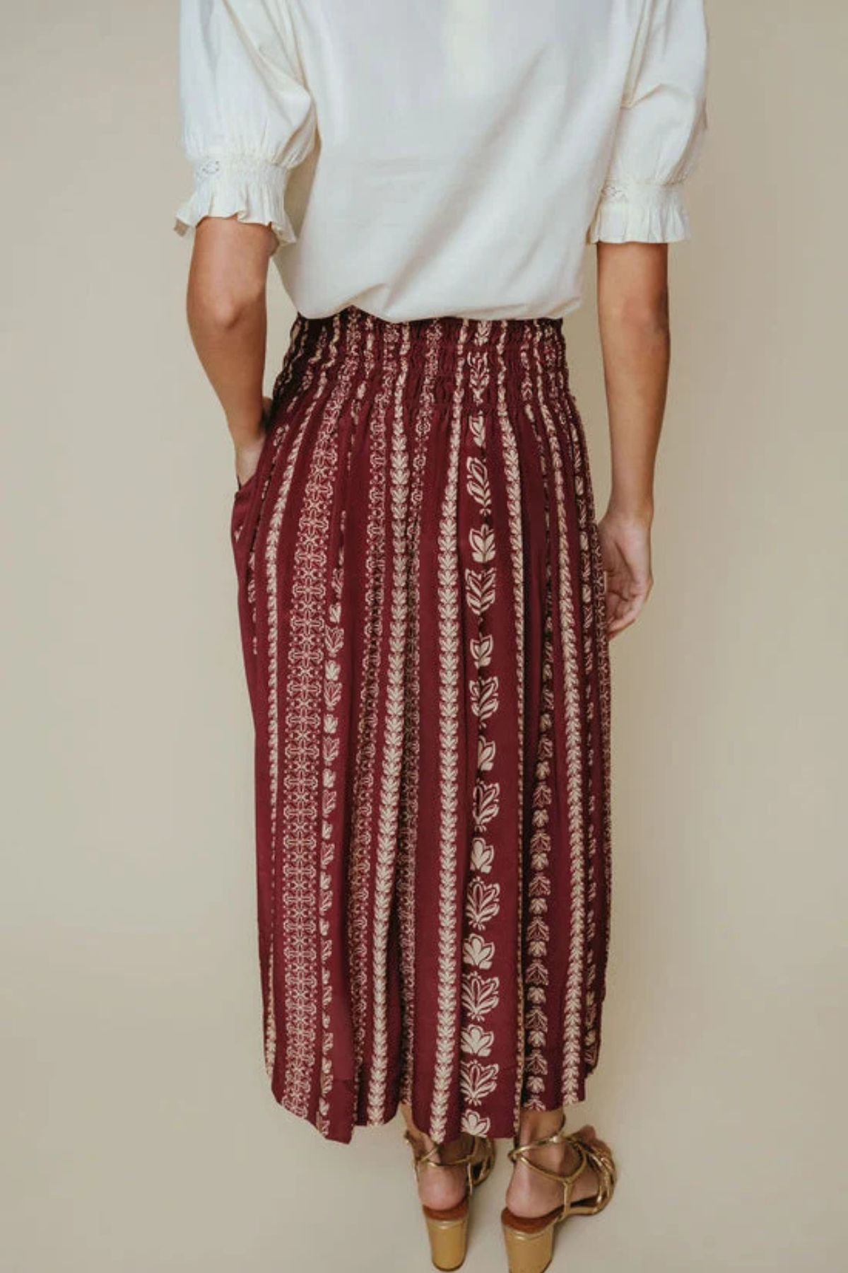 A person shown from the back wearing a white blouse and a high-waisted, pleated maxi skirt with vertical burgundy and beige leafy patterns, paired with brown heeled sandals against a solid beige background.