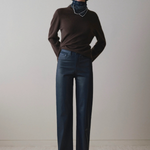 A model stands against a plain light beige wall wearing a dark brown long-sleeve turtleneck sweater, dark blue wide-leg jeans, and black pointed shoes.
