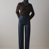 A model stands against a plain light beige wall wearing a dark brown long-sleeve turtleneck sweater, dark blue wide-leg jeans, and black pointed shoes.
